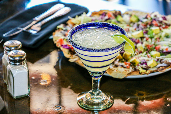 Un bicchiere di margarita su un tavolo di legno, accompagnato da un piatto di nachos e posate sullo sfondo