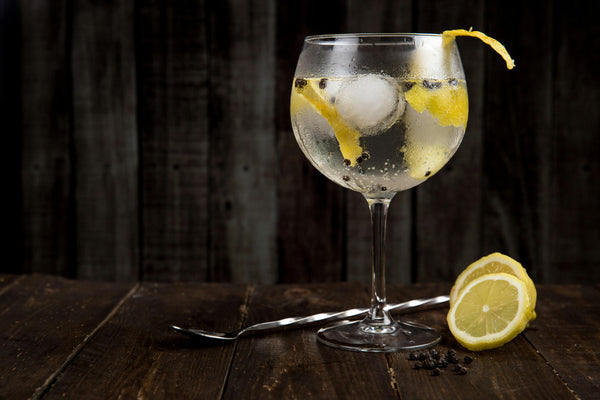 Bicchiere con Gin Tonic, ghiaccio, scorze di limone e bacche di ginepro, appoggiato su un tavolo di legno scuro con una fetta di limone accanto e un cucchiaino lungo