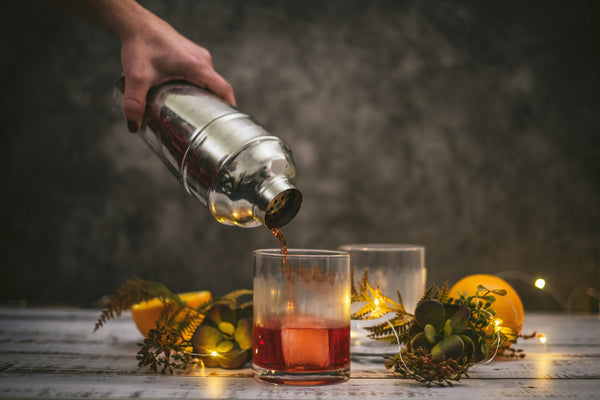 Una mano che versa un Negroni da uno shaker d'acciaio in un bicchiere di vetro contenente ghiaccio, posizionato su un tavolo decorato con elementi naturali come piante, agrumi e luci calde